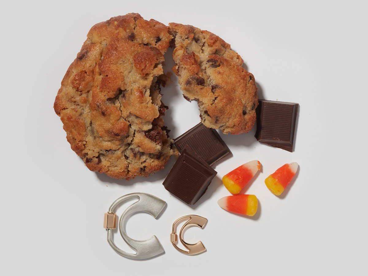 Large silver letter C charm and small rose gold letter C charm alongside a cookie, some pieces of chocolate, and some pieces of candy corn