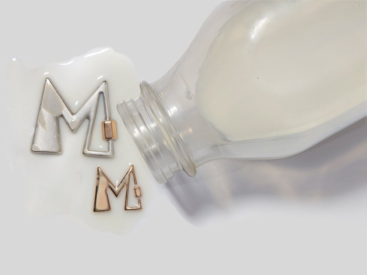Large silver letter M charm and small rose gold letter M charm with milk being poured on top of them