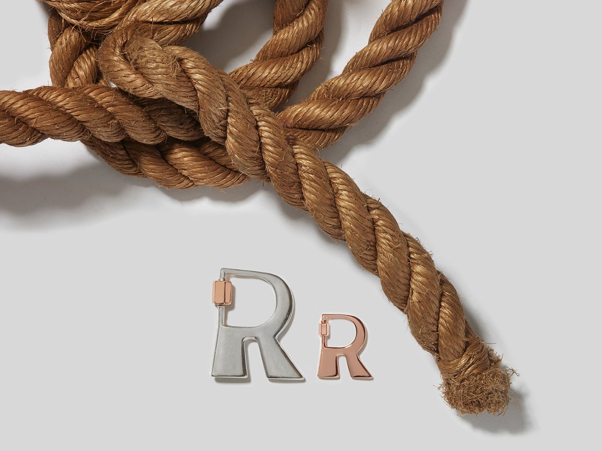 Big silver letter R charm and small rose gold letter R charm alongside knotted rope against white backdrop