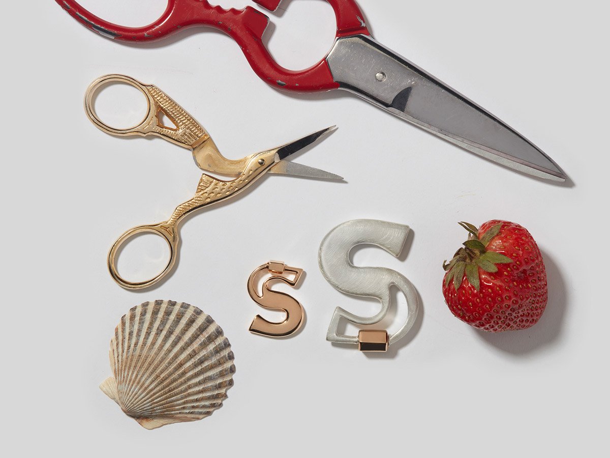 Big silver letter S charm and small rose gold letter S charm alongside strawberry, shell, and two pairs of scissors against white backdrop