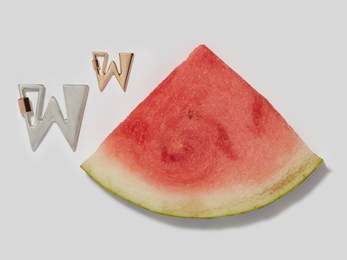 Big silver letter W charm and small rose gold letter W charm alongside slice of watermelon against white backdrop
