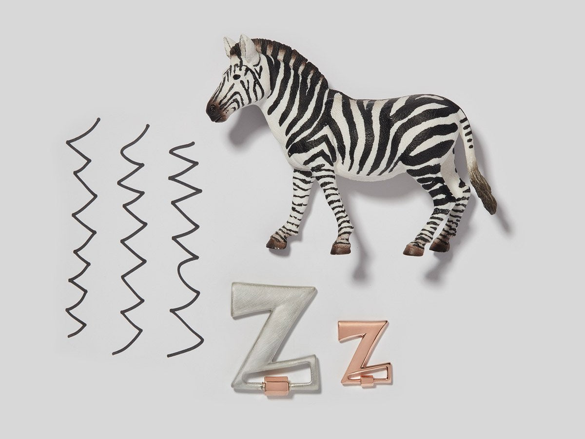 Big silver letter Z charm alongside small rose gold letter Z charm with zebra figurine and black zig zag lines against a white backdrop