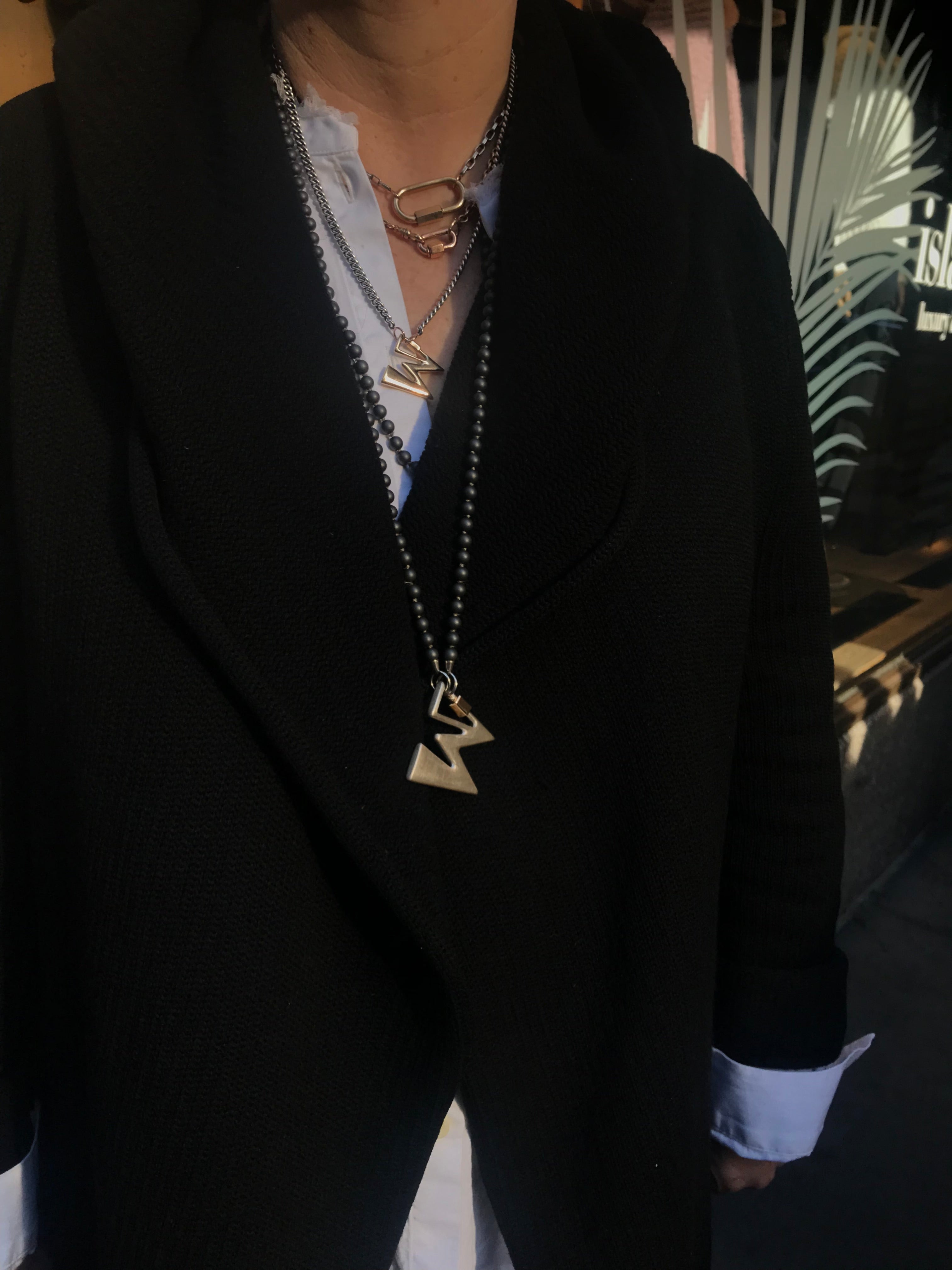 Close up of woman's torso wearing black trench coat and two long necklaces with W lock charms