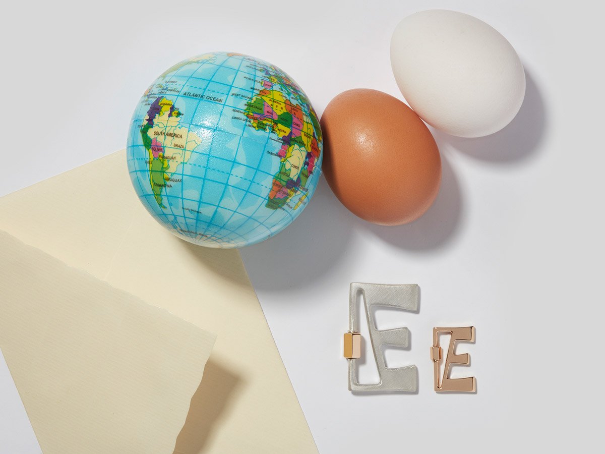 Large silver letter E charm and small rose gold letter E charm alongside a an envelope, two eggs, and small globe