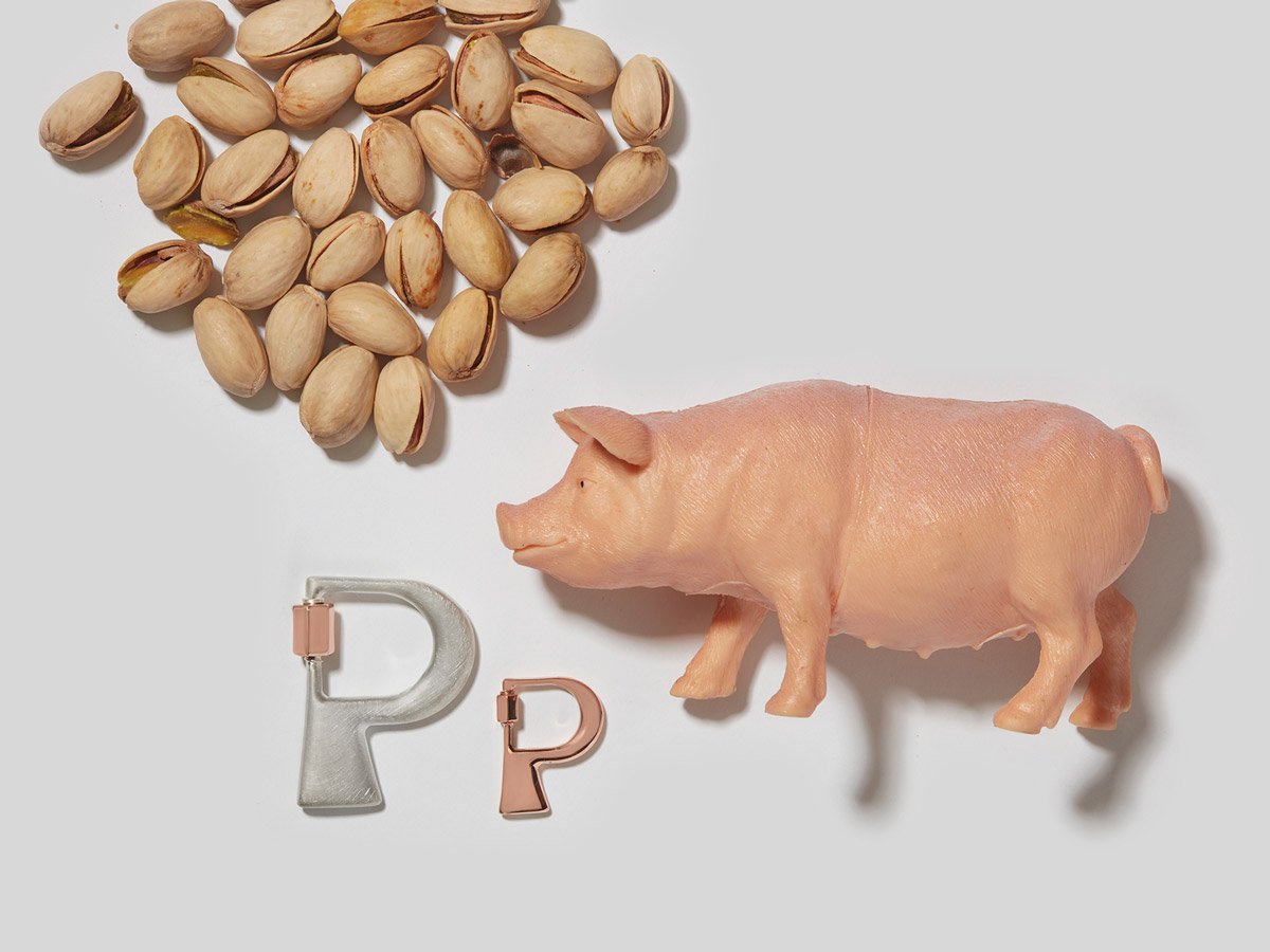 Large silver letter P charm and small rose gold letter P charm alongside pile of pistachios and a pig figurine