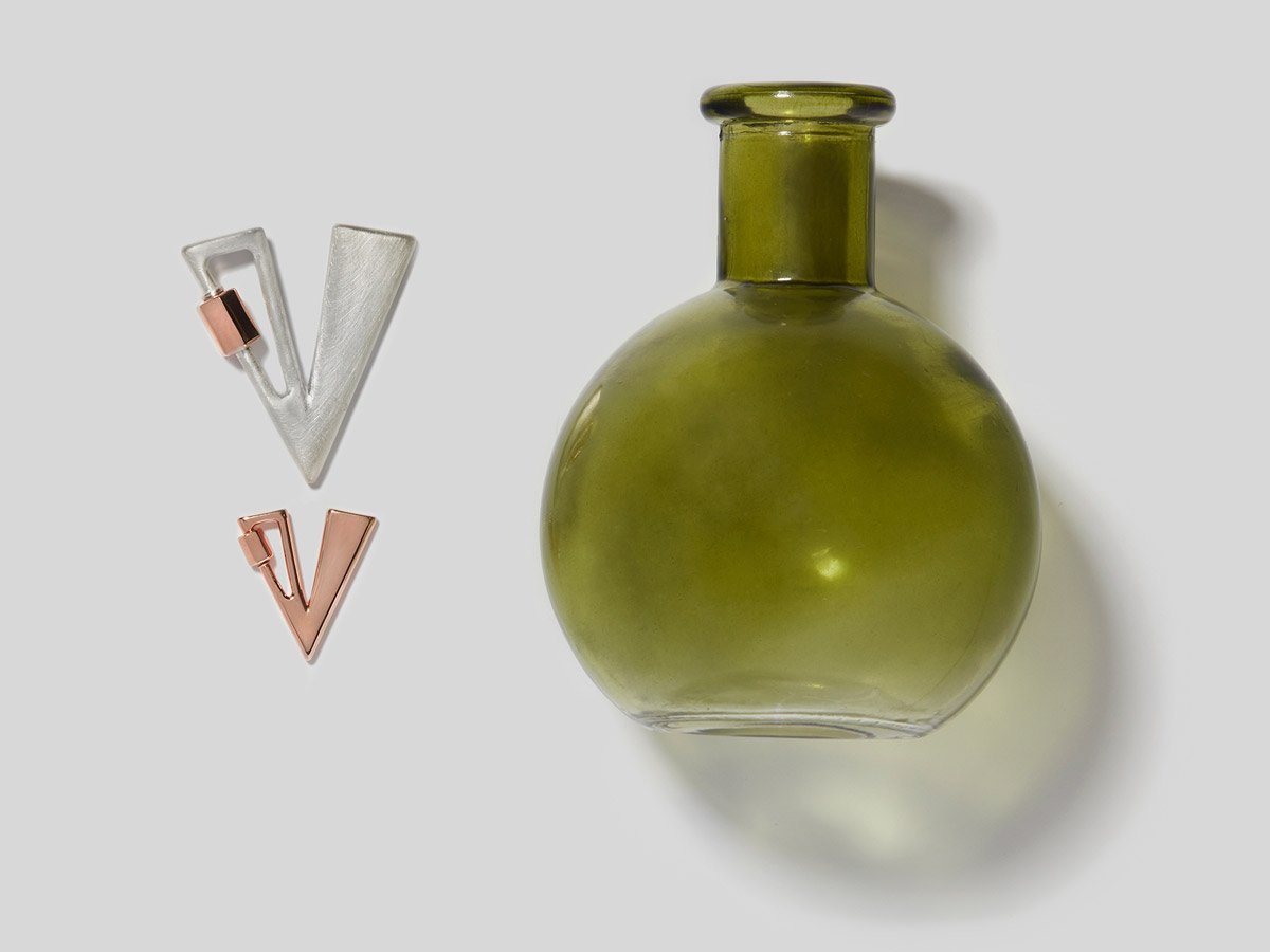 Big silver letter V charm and small rose gold letter V charm alongside small green circular vase  against white backdrop