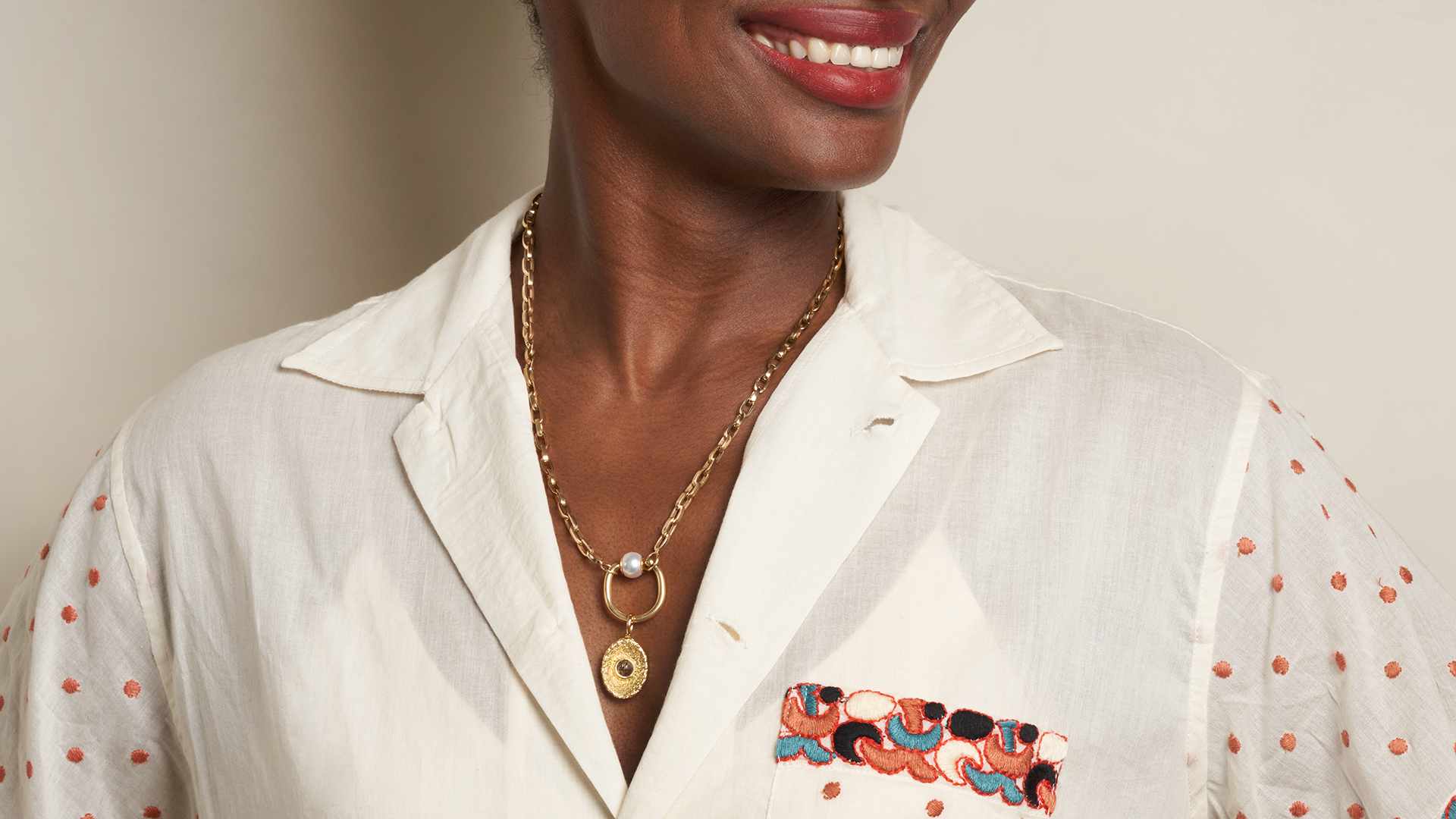 Close up of woman's decolletage wearing white embroidered shirt and gold necklace with trundle pearl ring